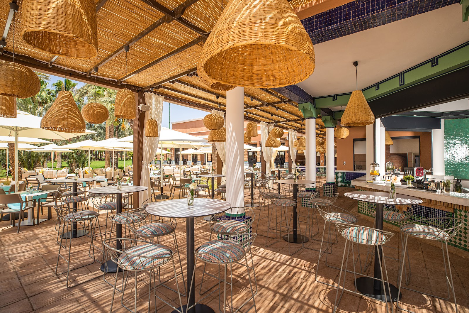 Covered dining terrace with woven pendant lights, round tables, and striped chairs at Sol Oasis Marrakech, overlooking palm-filled grounds and outdoor seating areas.