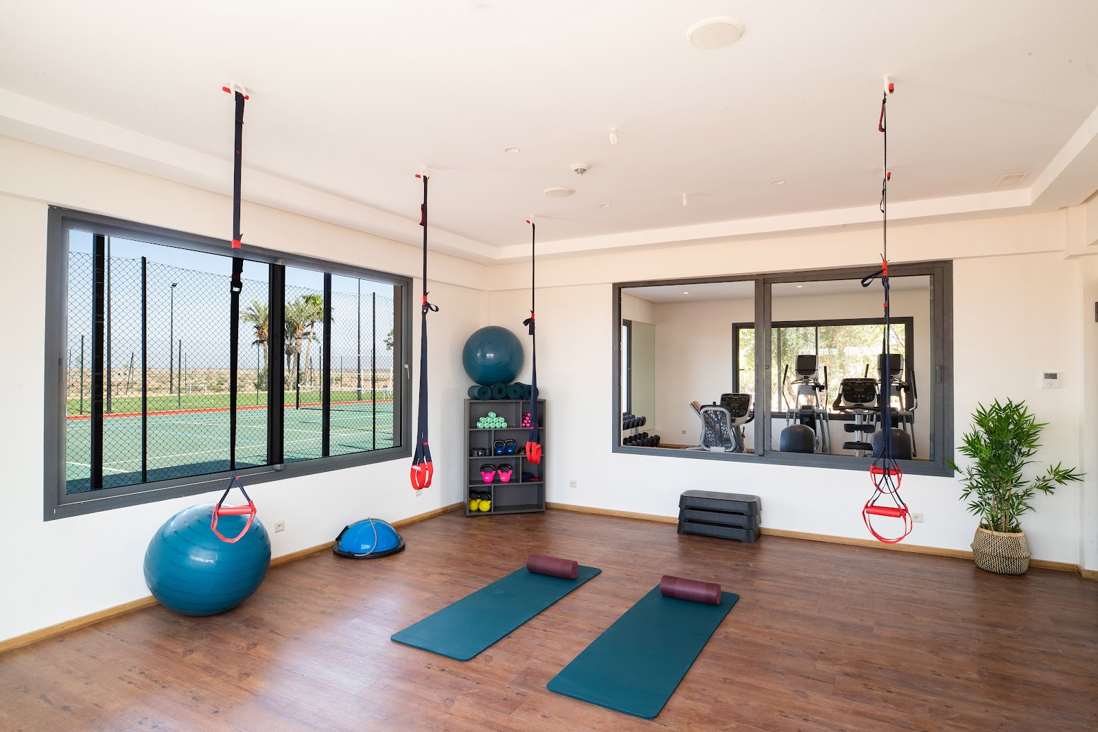 Resort fitness studio with yoga mats, exercise balls, suspended red gym rings, and a view of tennis courts through large windows.