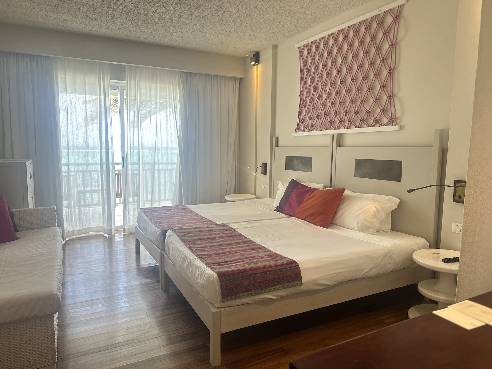 Resort guest room with queen bed, burgundy runner, decorative pillows, cream headboard, and glass doors opening to a balcony overlooking the ocean.