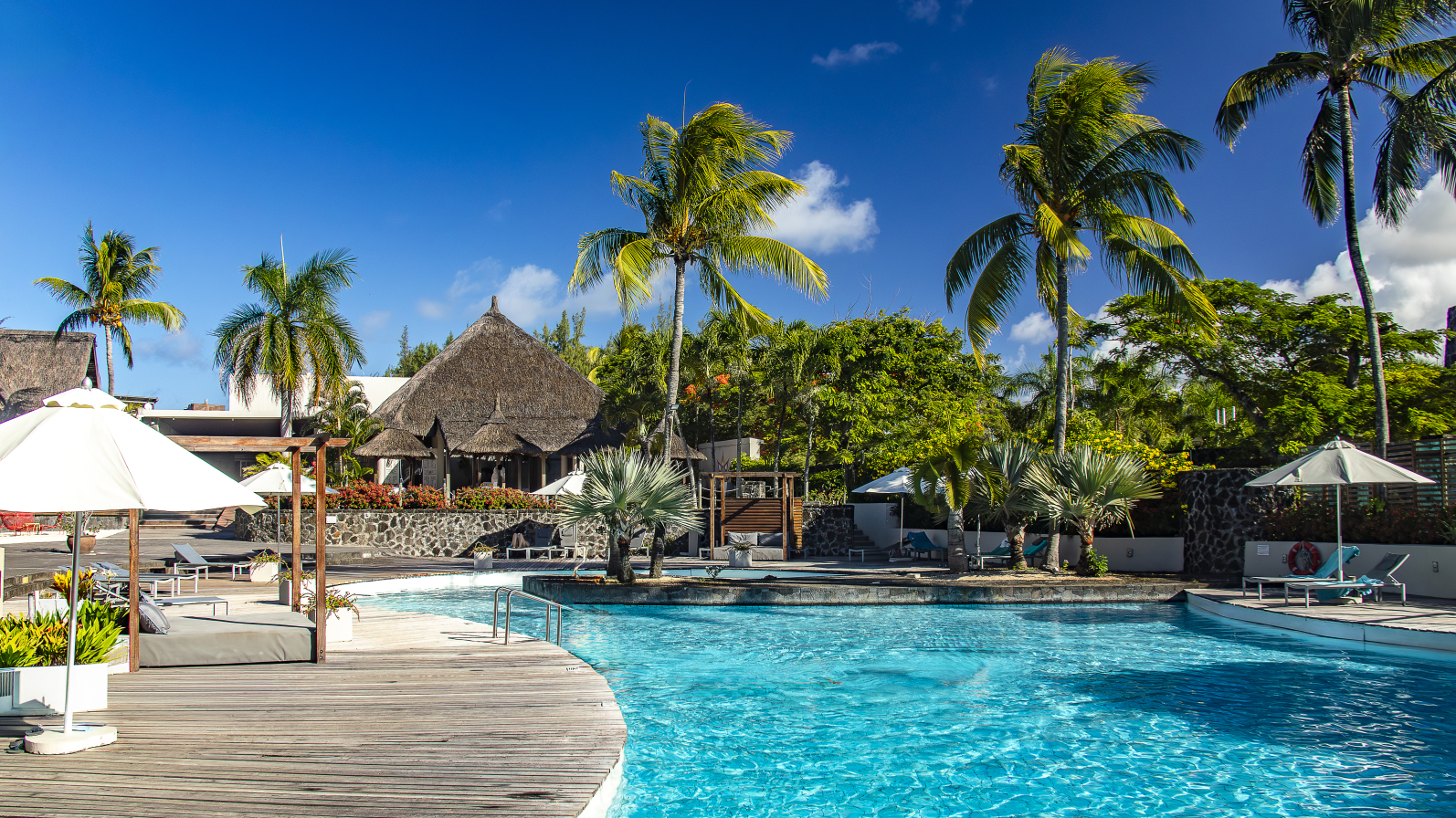 Wooden deck and bright turquoise pool at Solana Beach Mauritius, with thatched-roof bungalows, palm trees, and white umbrellas under clear blue sky.
