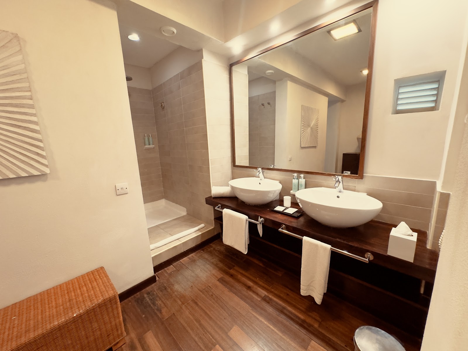 Modern bathroom with dual white vessel sinks on dark wood vanity, large mirror, walk-in shower, and wood-plank flooring.