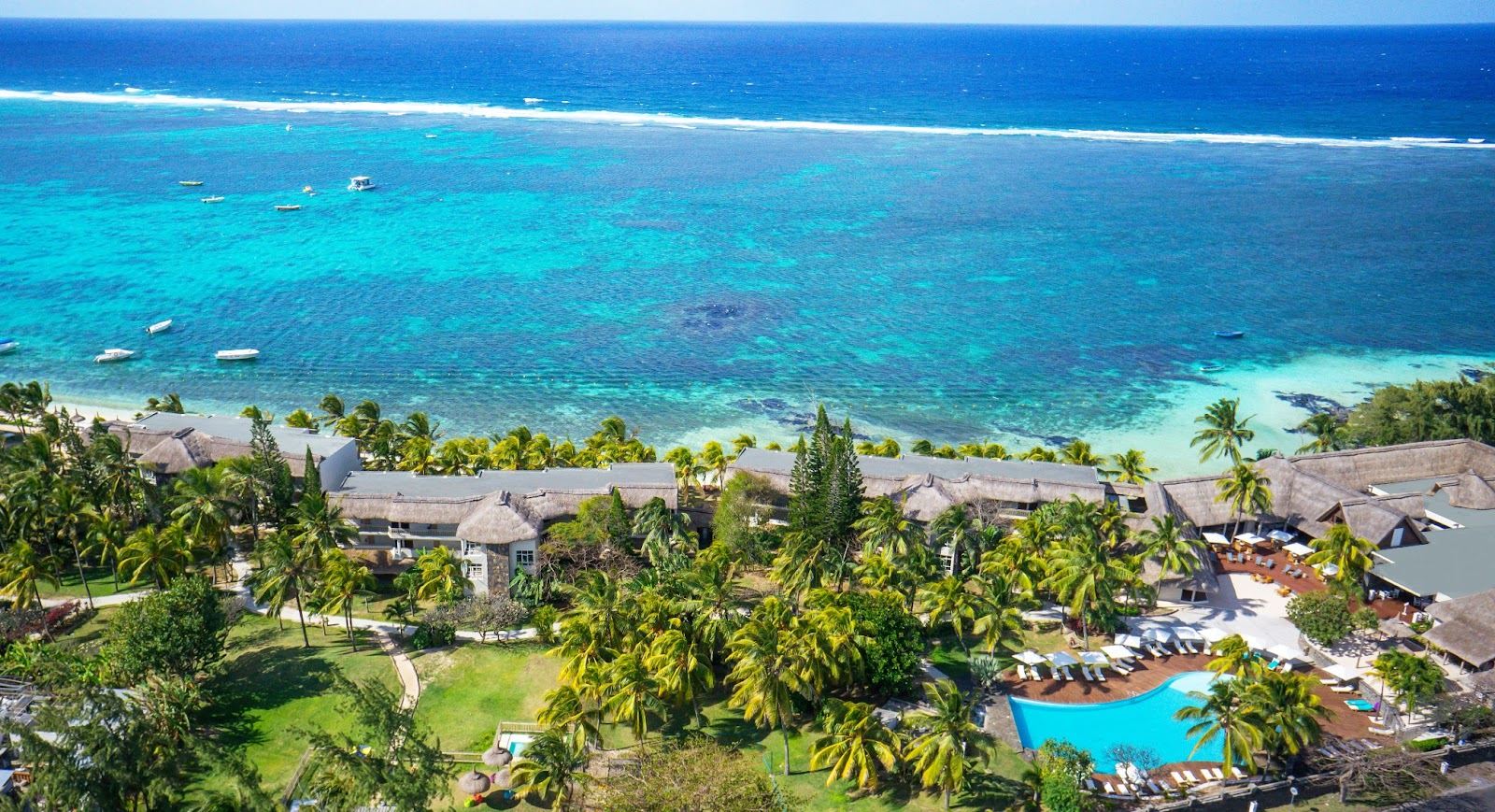 Aerial view of Solana Beach Mauritius with thatched bungalows, palm trees, blue pool, and turquoise lagoon with moored boats.
