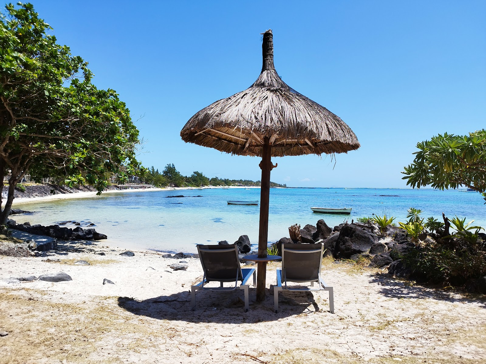 Two lounge chairs sit beneath a thatched umbrella on white sand beach, with turquoise water and moored boats beyond at Solana Beach Mauritius.