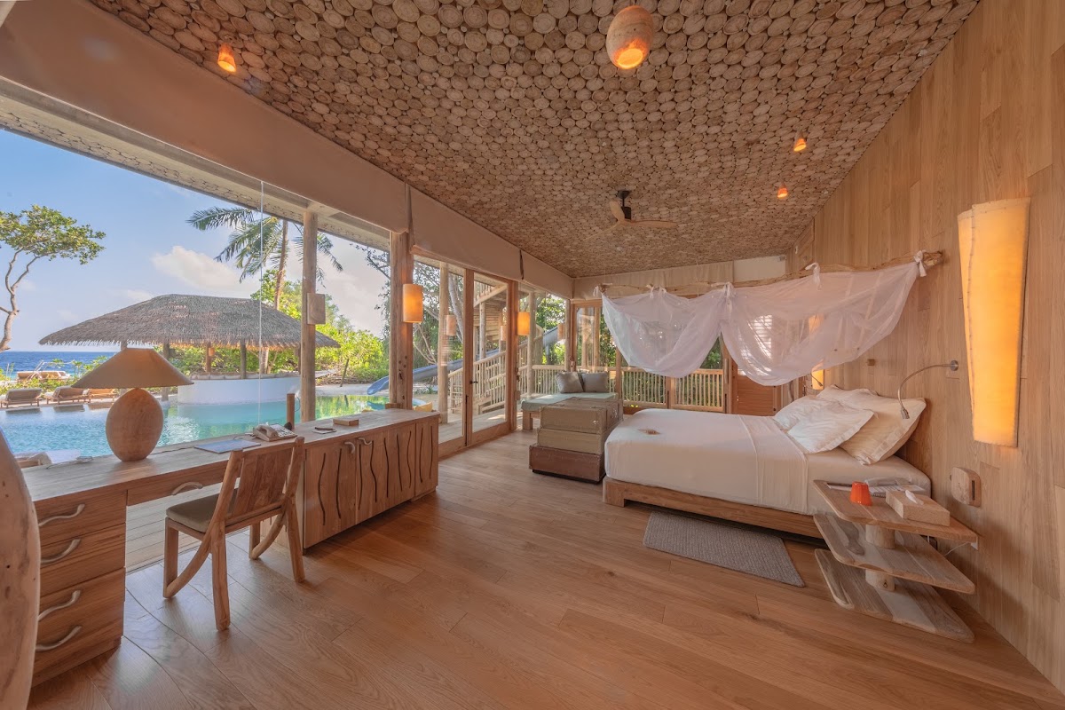 Overwater villa bedroom with wooden finishes, bed with sheer canopy, and ocean views through floor-to-ceiling windows at Soneva Fushi.