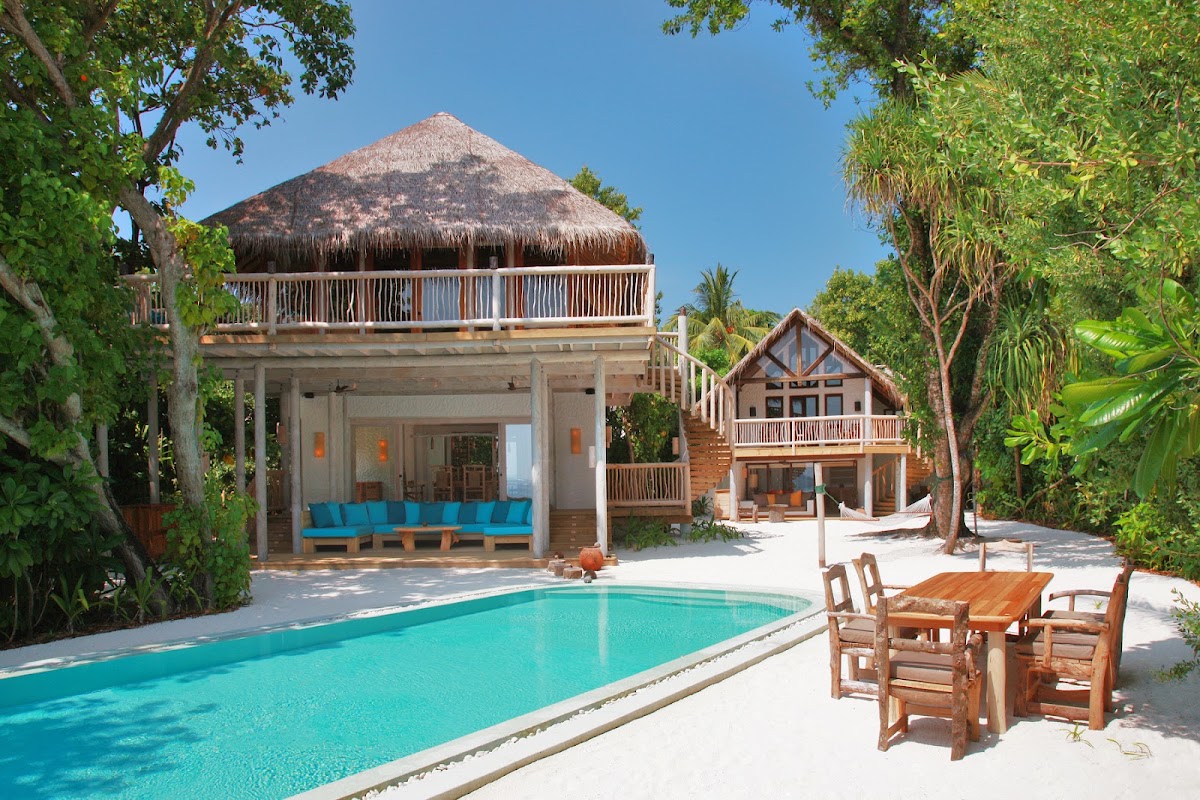 Turquoise pool with wooden dining table beside thatched-roof villas surrounded by tropical palms at Soneva Fushi.