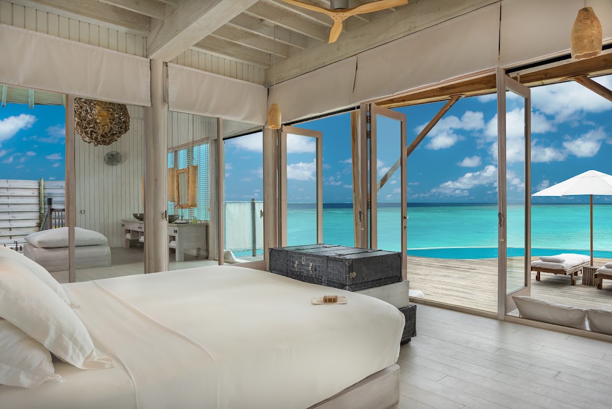 Overwater bungalow bedroom at Soneva Fushi with turquoise lagoon views through glass doors and wooden deck with loungers.
