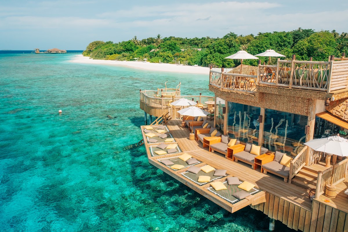 Wooden overwater bungalows with loungers and umbrellas extend into turquoise lagoon, with white sand beach and tropical vegetation visible in the distance.