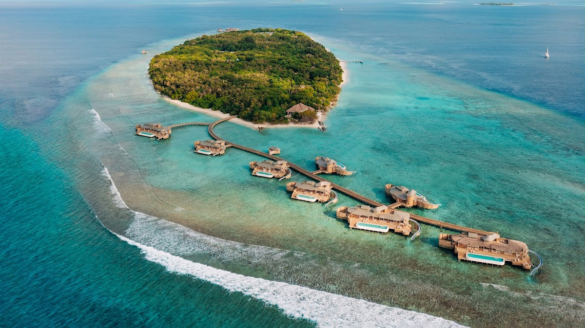 Aerial view of Soneva Fushi showing overwater bungalows connected by wooden walkways, with a forested island and turquoise lagoon.