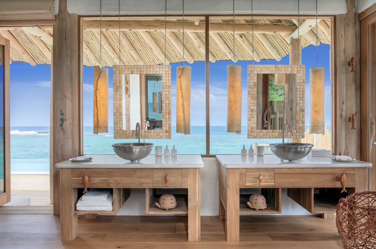 Soneva Fushi bathroom with dual wooden vanities, stone vessel sinks, and ocean views through open windows framing turquoise water and beach.