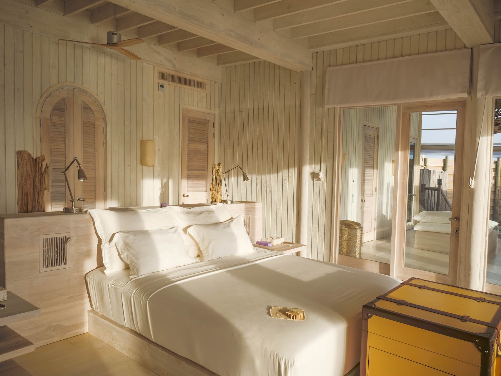 Light-filled bedroom with cream wood paneling, white bedding, and glass doors opening to a beachfront deck at Soneva Jani.