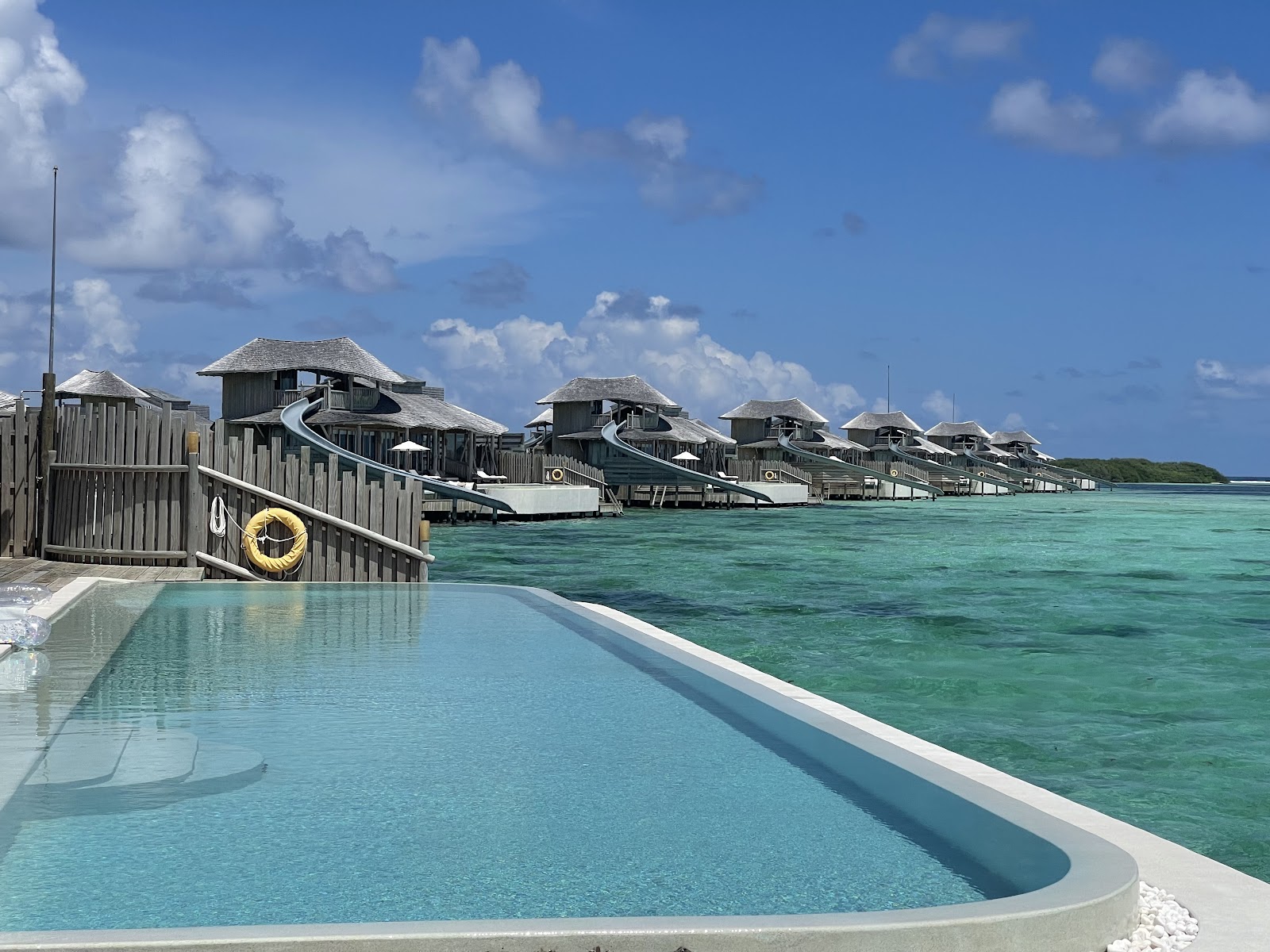 Curved pool with thatched-roof overwater bungalows and water slides stretching across turquoise lagoon at Soneva Jani.