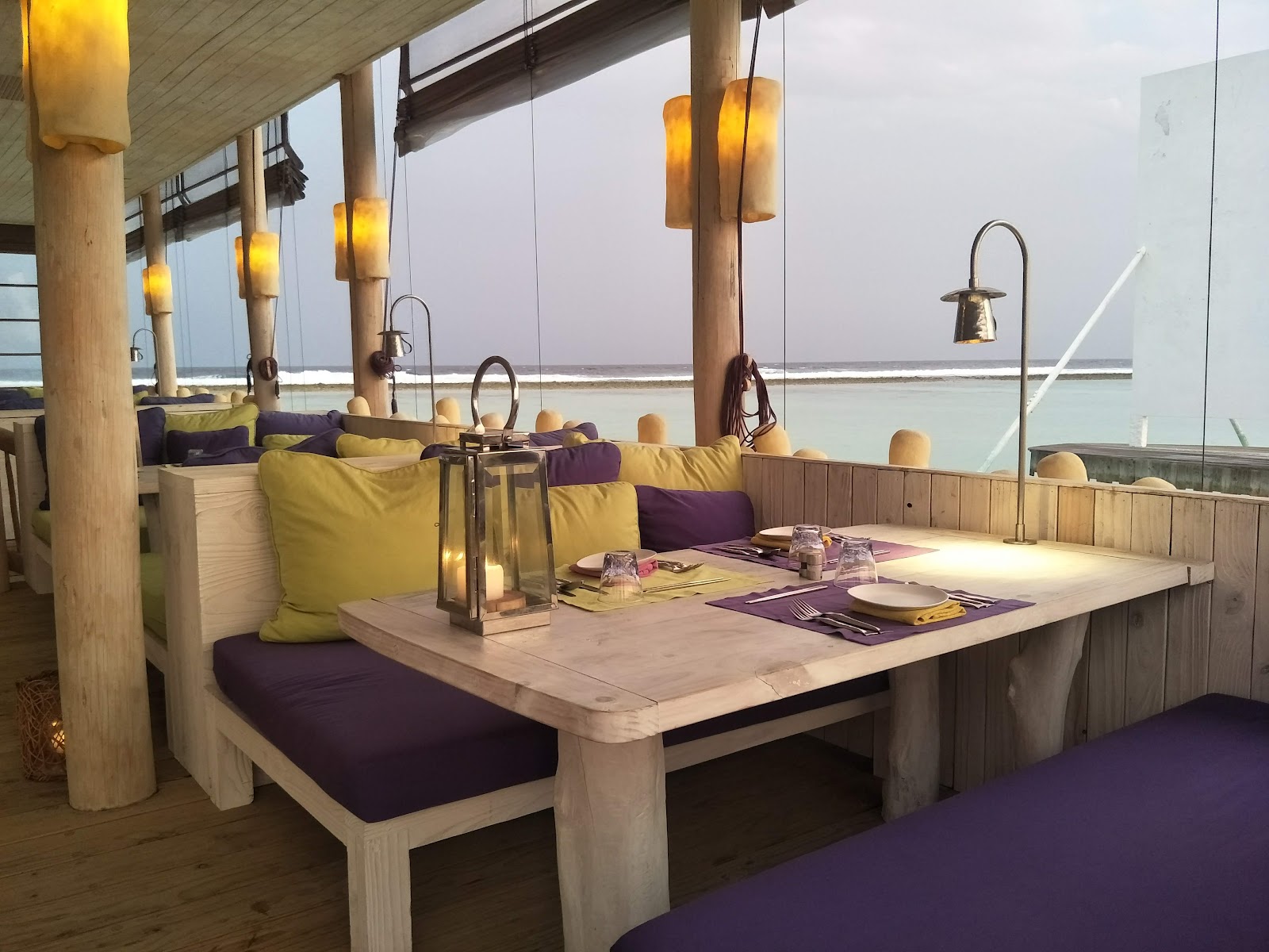 Soneva Jani's beachfront dining pavilion features wooden tables with purple and yellow cushioned seating, hanging amber lanterns, and ocean views through glass panels.