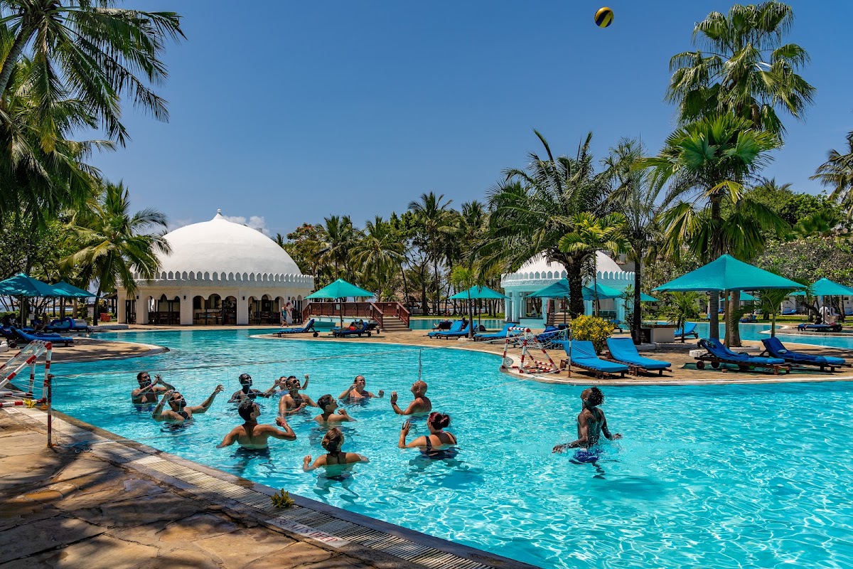 Guests swim and play volleyball in the turquoise pool at Southern Palms Beach Resort, surrounded by palm trees, blue sun loungers, and white domed structures.