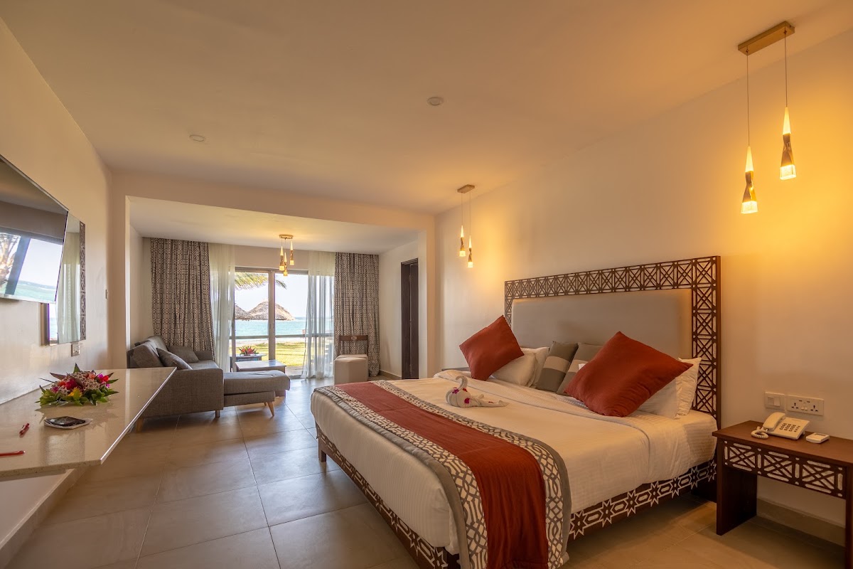 Spacious resort bedroom with king bed, red accent pillows, and ocean view through glass doors leading to balcony.