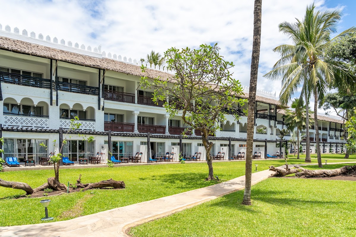 White colonial-style resort building with thatched roof overlooking manicured lawn, palm trees, and beachfront lounge chairs.