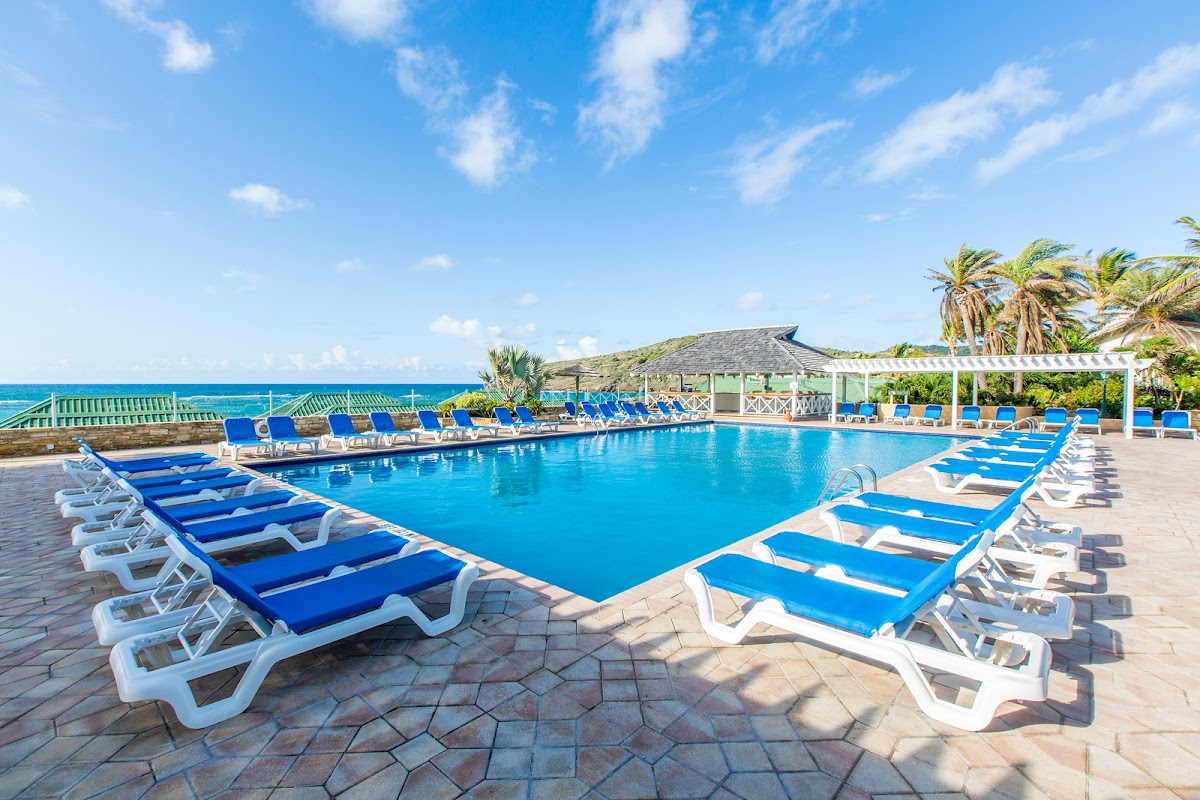 Resort pool surrounded by blue lounge chairs with ocean and palm trees visible beyond under clear sky.