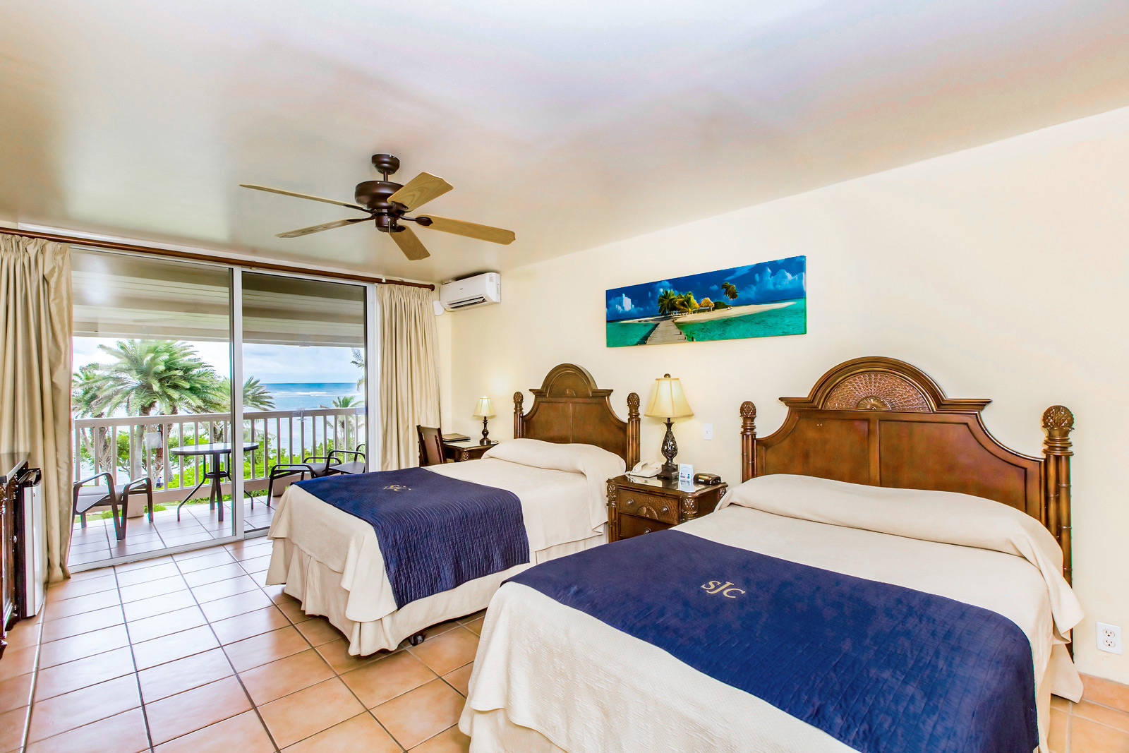 Resort guest room with two queen beds, wooden furnishings, and a balcony overlooking the ocean and palm trees.