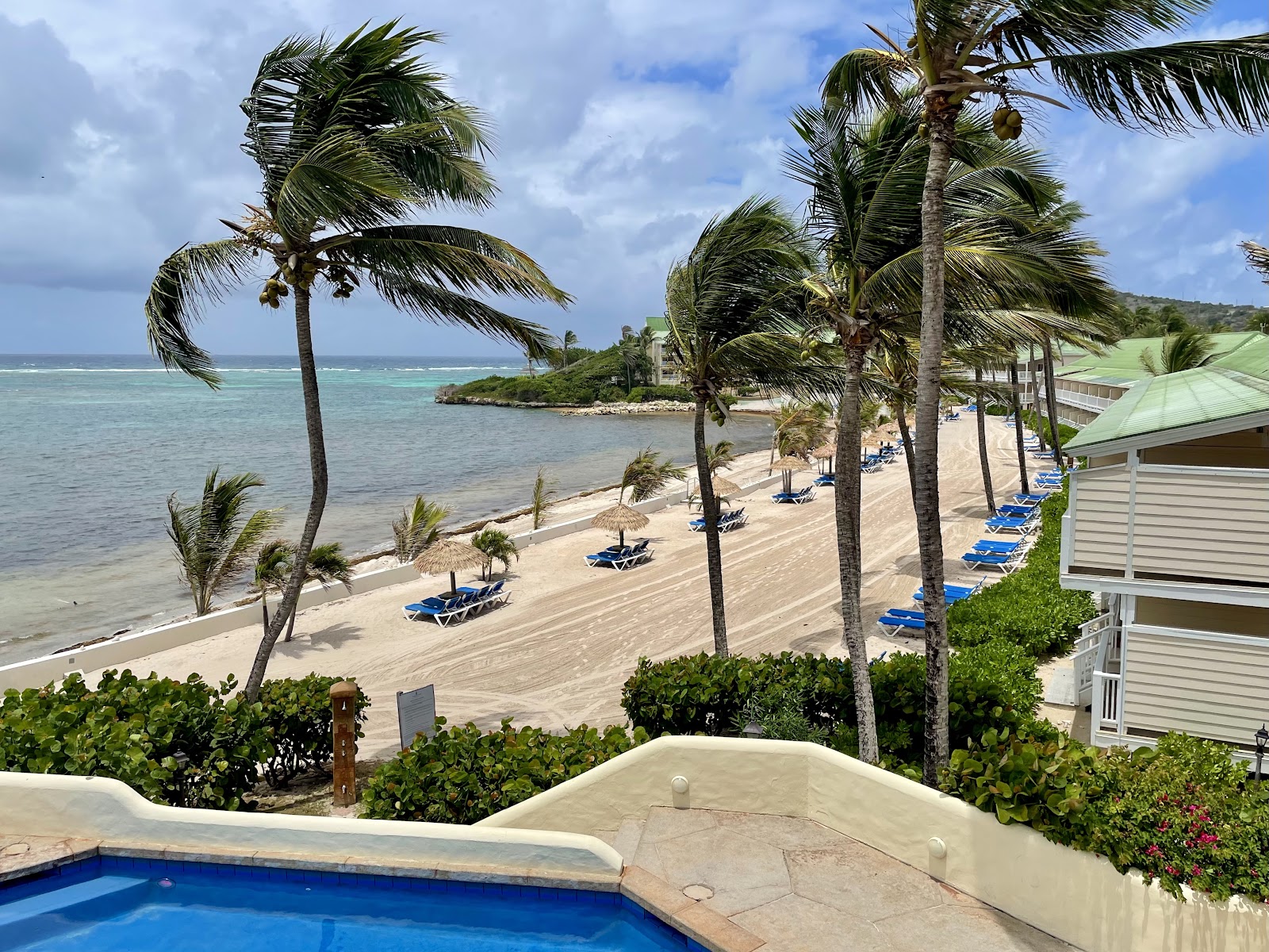Pool deck and beach at St. James's Club & Villas with palm trees, blue loungers on sand, and turquoise Caribbean waters beyond.