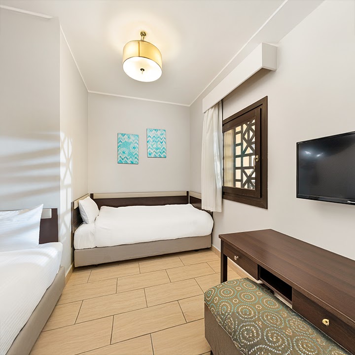 Resort hotel bedroom with two beds, wooden furniture, wall-mounted TV, and turquoise artwork.