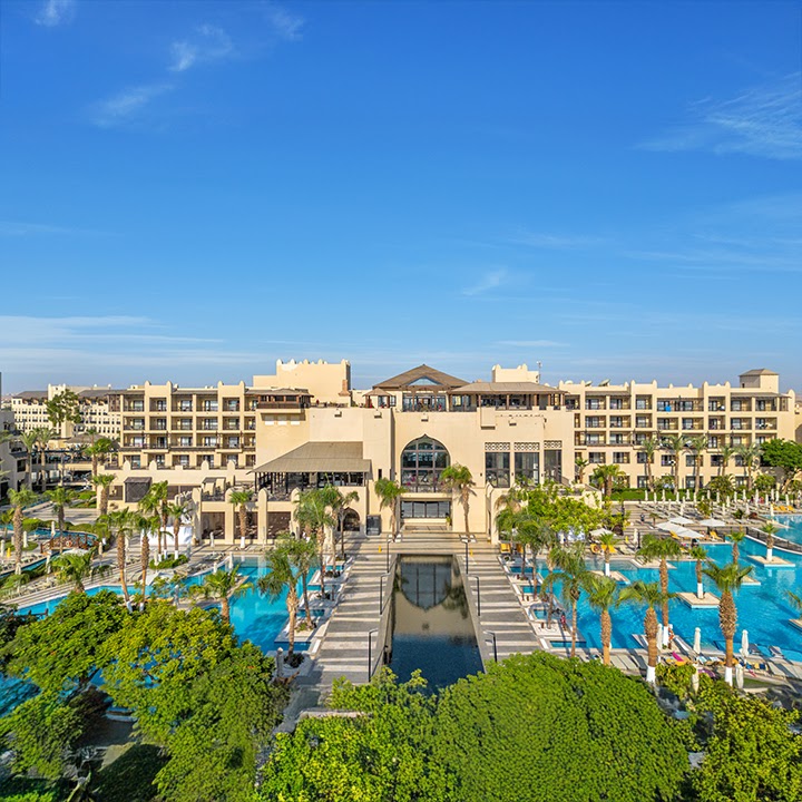 Steigenberger Aqua Magic Hotel's beige buildings overlook multiple blue pools, palm trees, and a central water feature under clear sky.