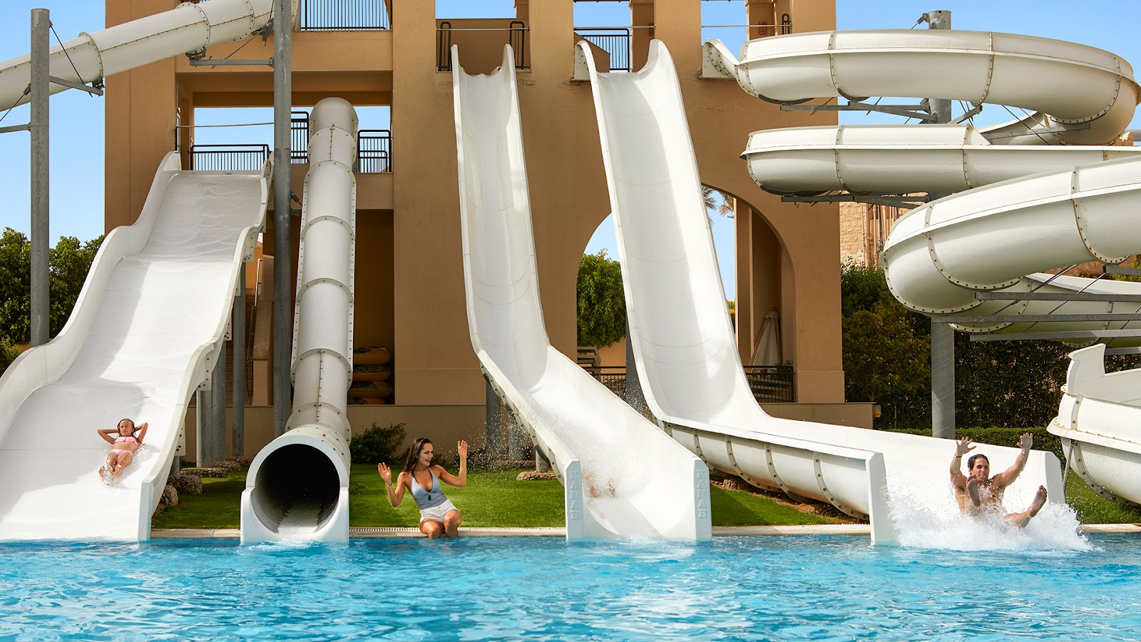 Large bright blue pool with white umbrellas and loungers, surrounded by palm trees and tan resort buildings under clear sky.