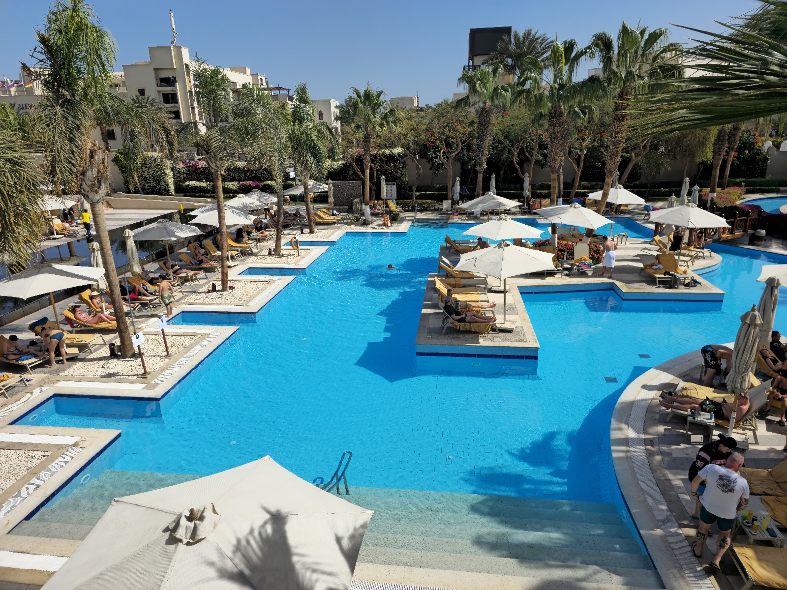 Large turquoise pool with island features and palm trees, surrounded by lounge chairs and cream umbrellas at Steigenberger Aqua Magic Hotel.