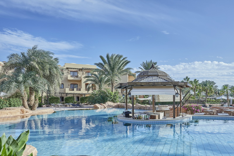Large blue pool with thatched bar pavilion, palm trees, and sandy-colored resort buildings under clear sky at Steigenberger Coraya Beach.
