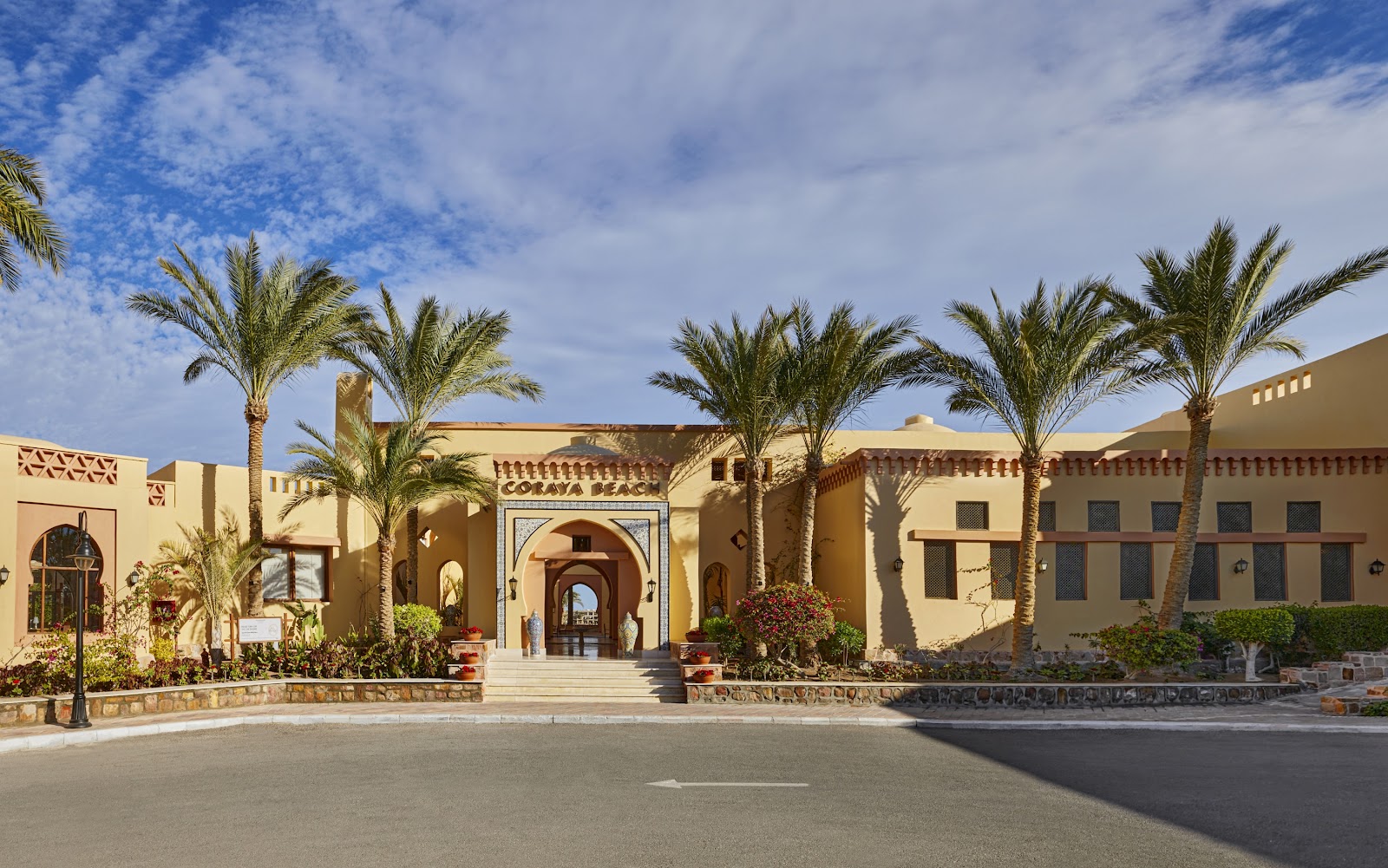 Tan stucco resort buildings with arched doorways and blue windows framed by tall palm trees under blue sky at Steigenberger Coraya Beach.