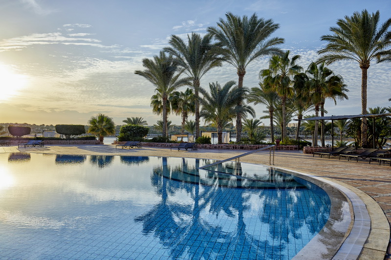 Resort pool with blue tiled floor surrounded by palm trees at sunrise, wooden deck, and lounge chairs in the distance.