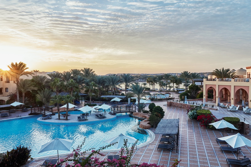 Curved pool with white umbrellas and loungers overlooking palm trees, the Nile River, and resort buildings at sunrise.