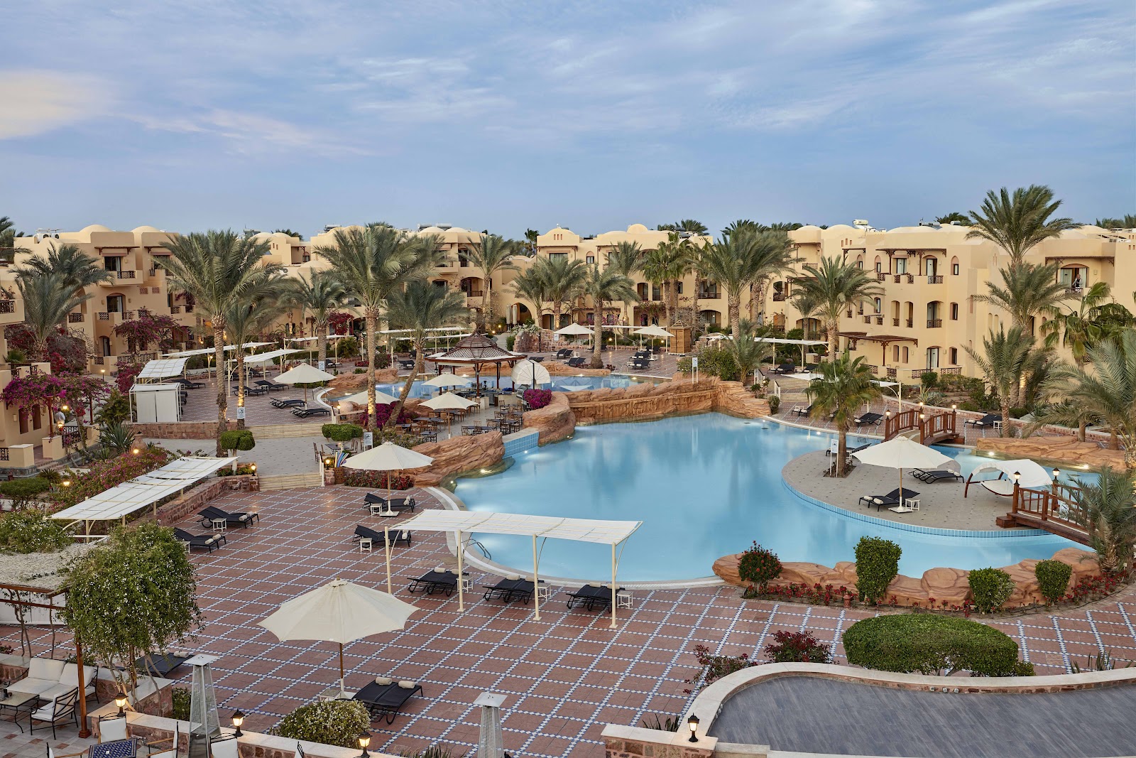 Large pool complex surrounded by cream-colored resort buildings, palm trees, and white umbrellas at Steigenberger Coraya Beach.