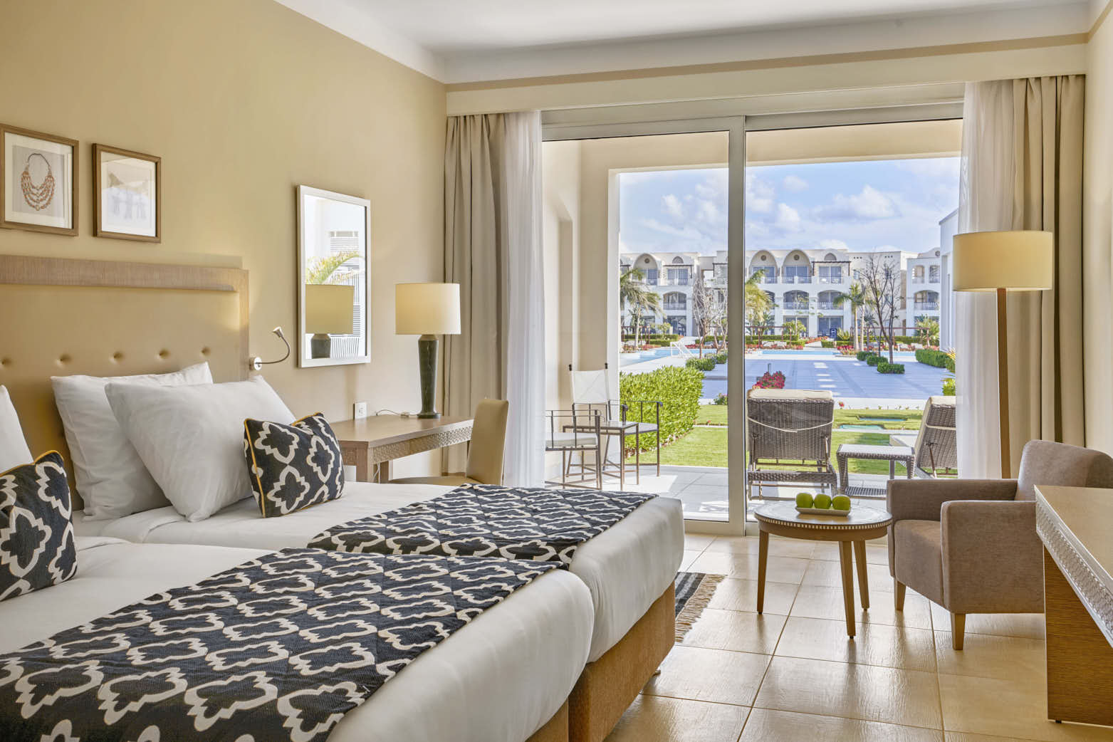 Spacious bedroom with cream walls, two beds with patterned runners, and glass doors opening to a courtyard with white-walled buildings and landscaped grounds at Steigenberger Resort Alaya Marsa Alam.