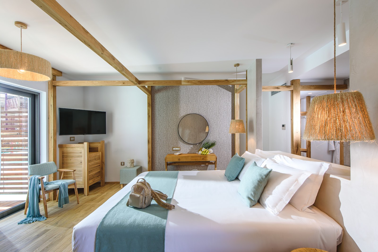 Resort suite with wooden beam ceiling, canopy bed with white linens and teal pillows, wall-mounted TV, and woven pendant lights.