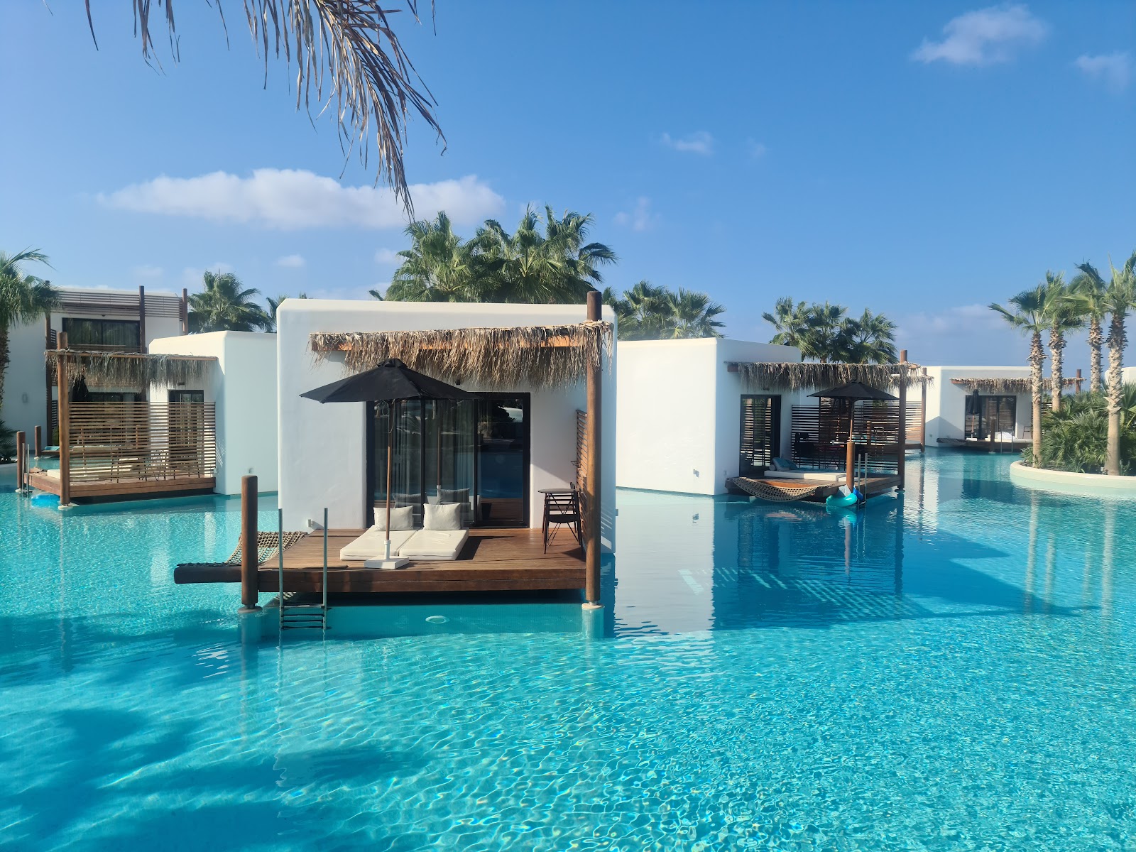 White overwater bungalows with thatched roofs line a turquoise lagoon at Stella Island Luxury Resort & Spa, with palm trees and clear sky beyond.