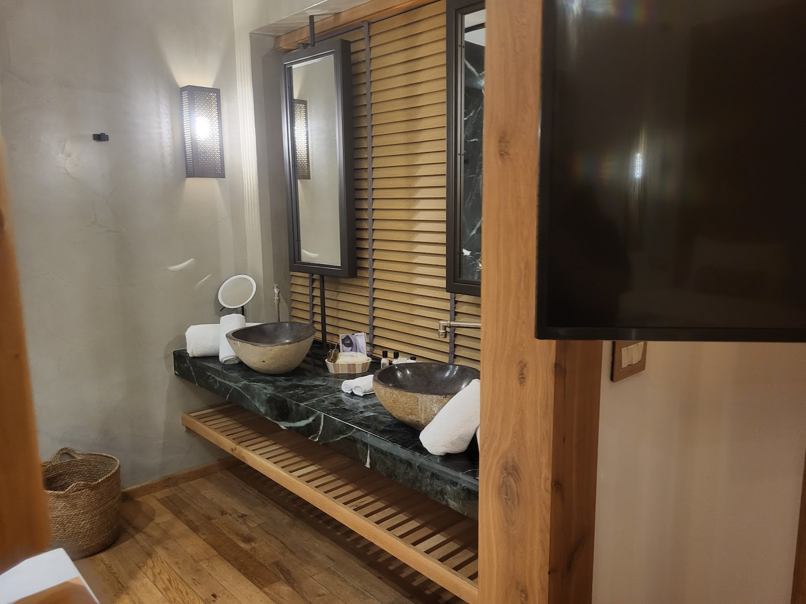 Resort bathroom with dual stone vessel sinks, wooden slat accents, and round mirrors above a dark marble countertop.