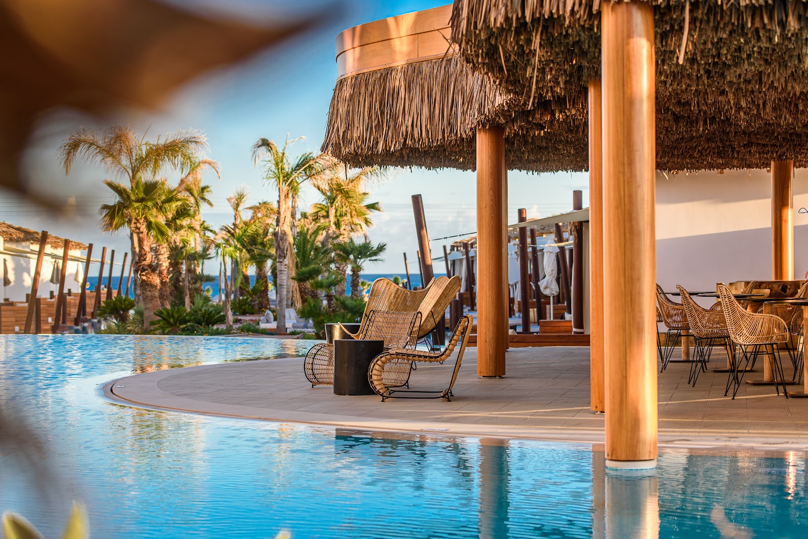 Pool deck at Stella Island with woven lounge chairs, thatched cabana, palm trees, and blue sea beyond under clear sky.