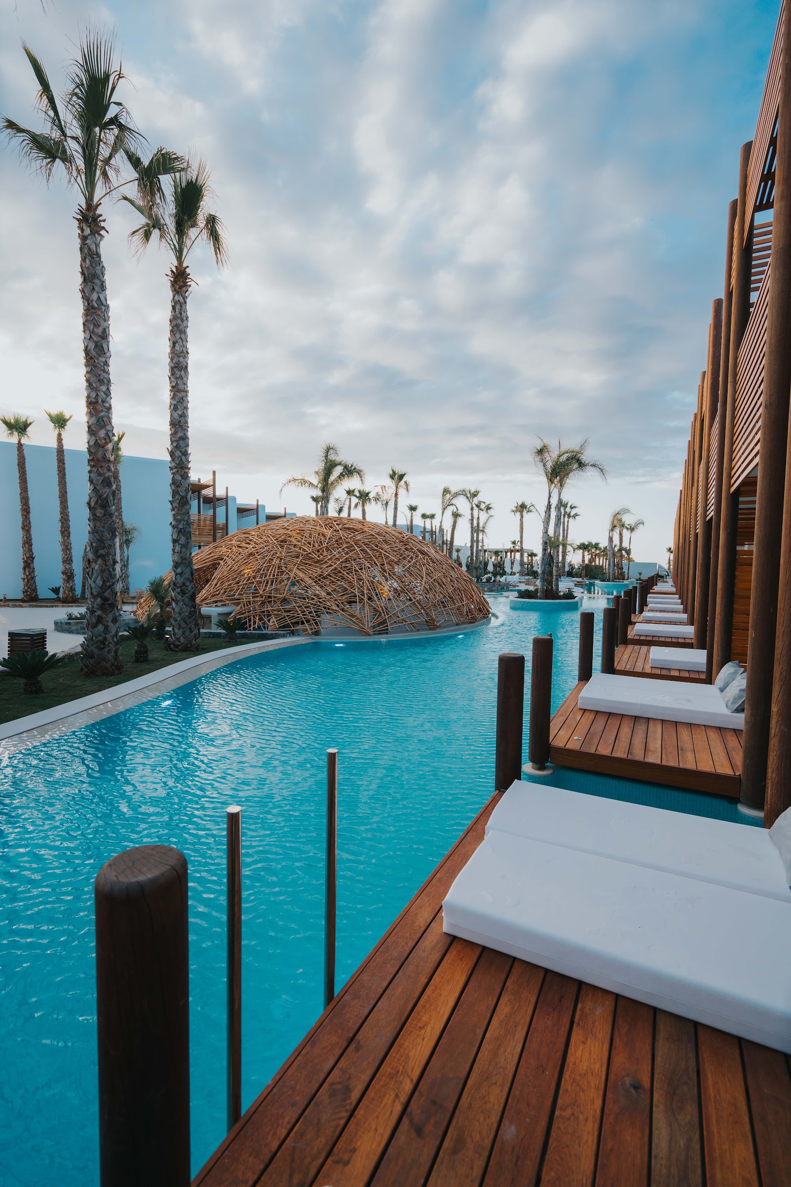 Wooden loungers line a turquoise pool with a woven dome structure, flanked by tall palms and modern brown buildings under cloudy sky.