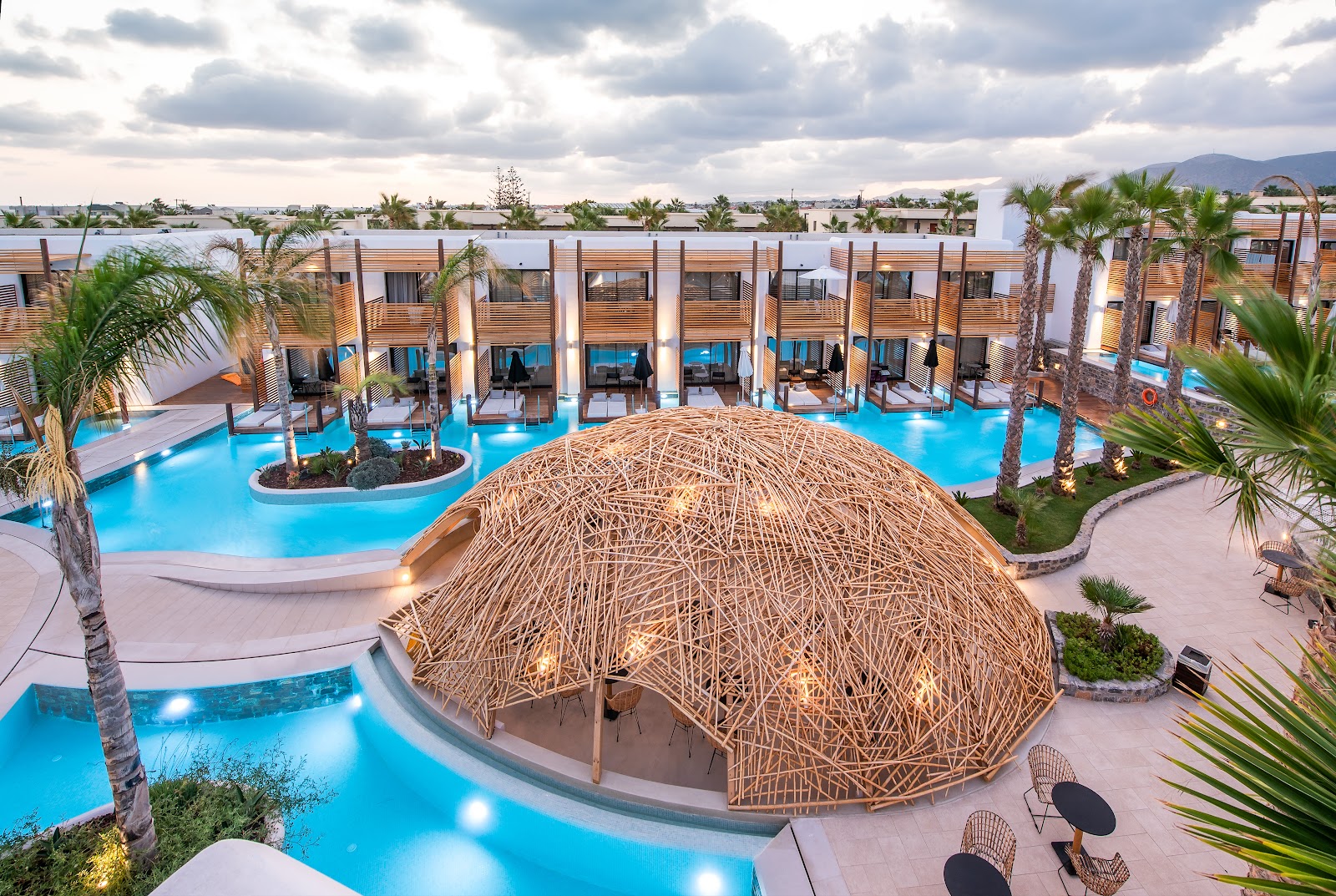 Stella Island Luxury Resort & Spa features a large woven dome structure on the pool deck surrounded by bright blue pools, palm trees, and modern white guest buildings.