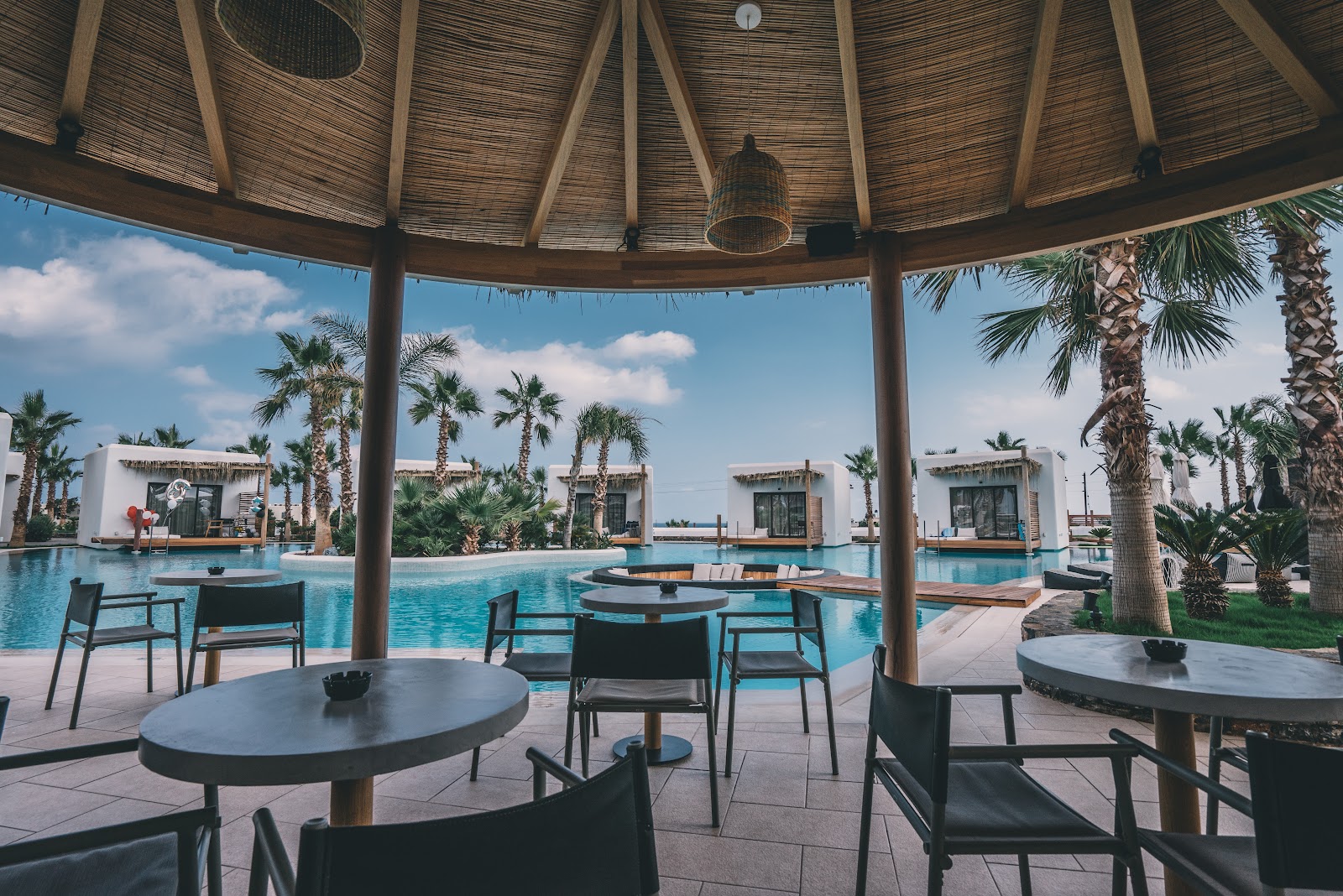 Illuminated thatched-roof bar structure with wooden columns overlooks glowing blue pools at Stella Island Luxury Resort at dusk.