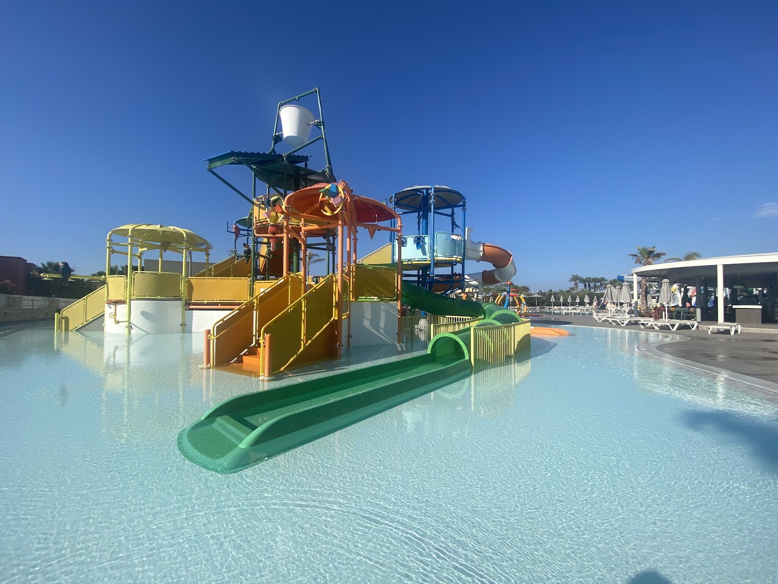 Colorful water play structures with slides and climbing features in a shallow turquoise pool at Stella Palace, with loungers and palm trees visible beyond.