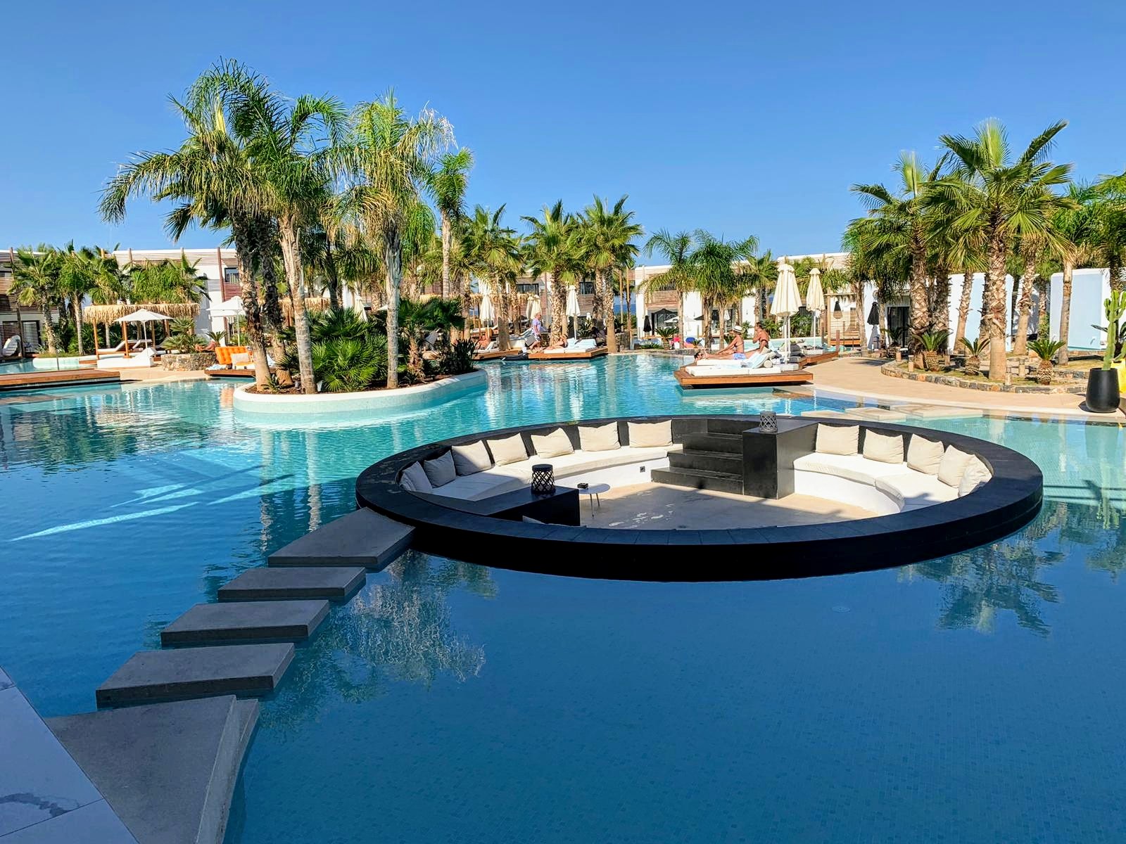 Circular sunken lounge platform in resort pool surrounded by palm trees, with beach cabanas and whitewashed buildings in background.