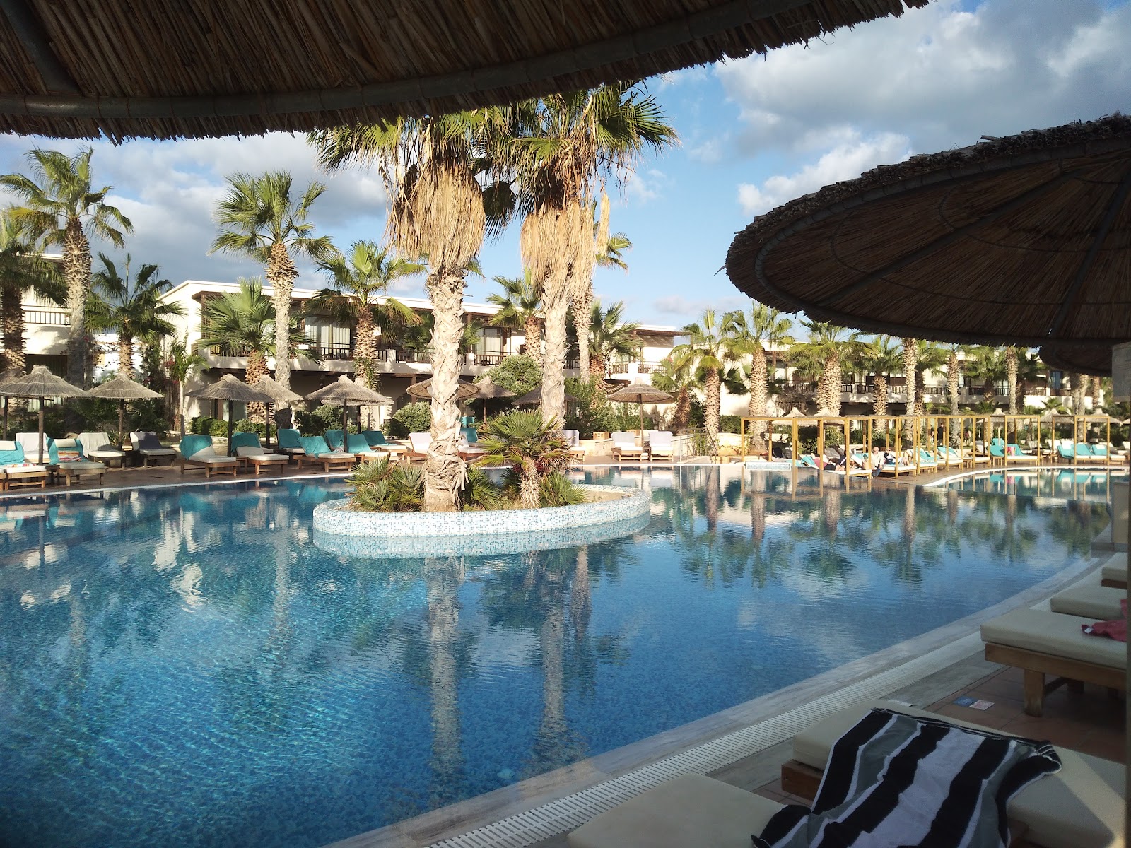 Large lagoon-style pool with tall palms and central island, shaded by thatched roof, resort buildings and loungers in background.
