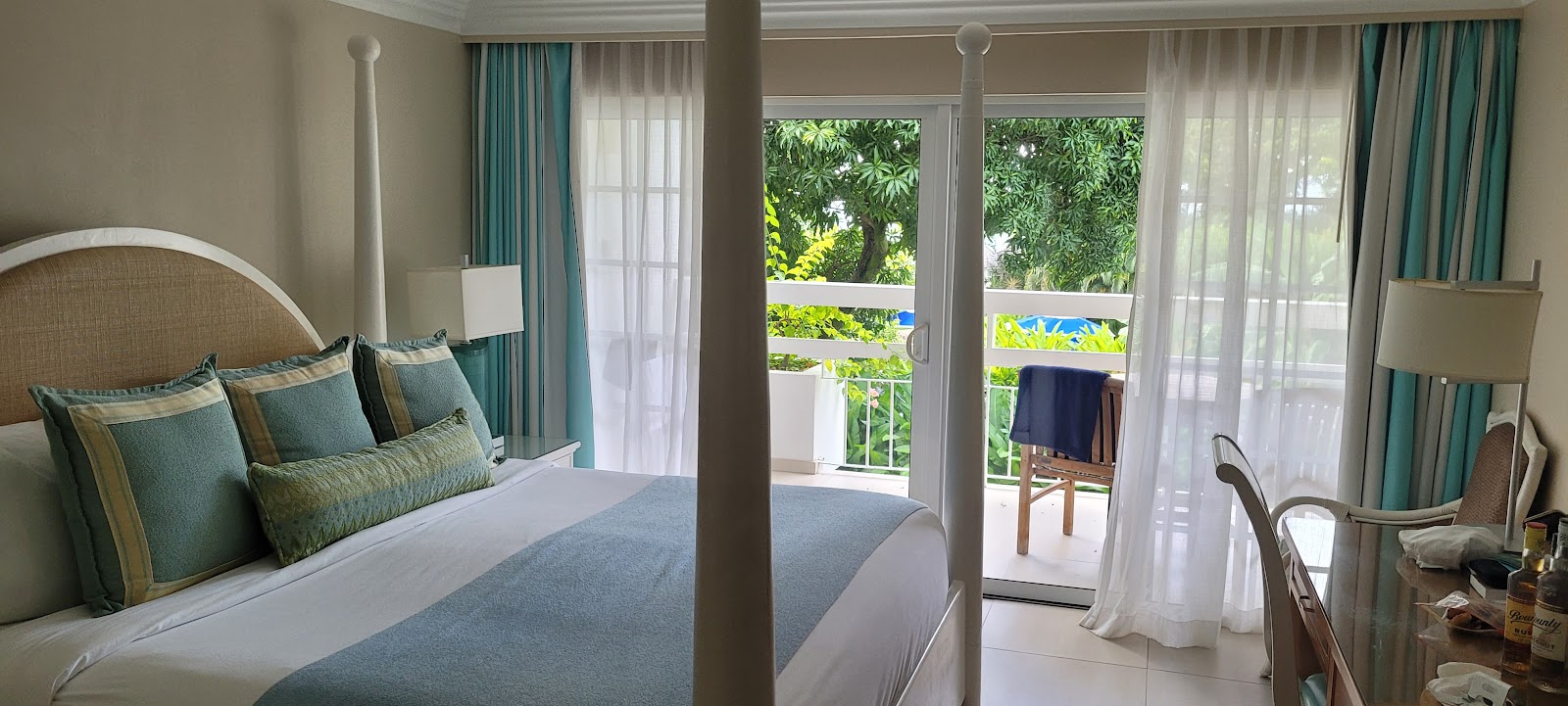 Resort bedroom with four-poster bed, turquoise pillows, and glass doors opening to a balcony overlooking tropical gardens and ocean.