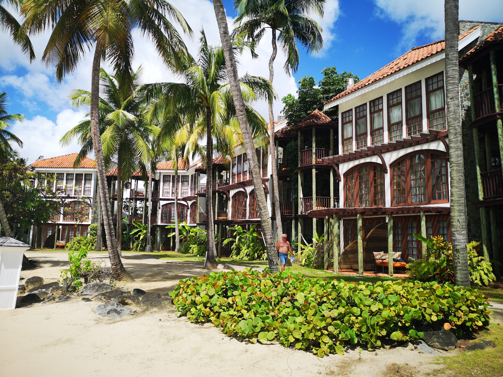 Colonial-style beachfront buildings with tile roofs, wooden balconies, and palm trees surrounding a sandy courtyard at StolenTime by Rendezvous.