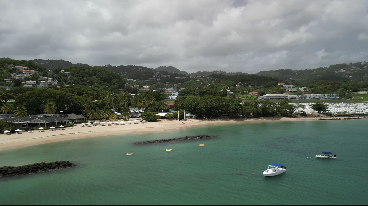 Sandy beach with turquoise water, moored boats, and palm trees at StolenTime by Rendezvous, with forested hills and resort buildings beyond.