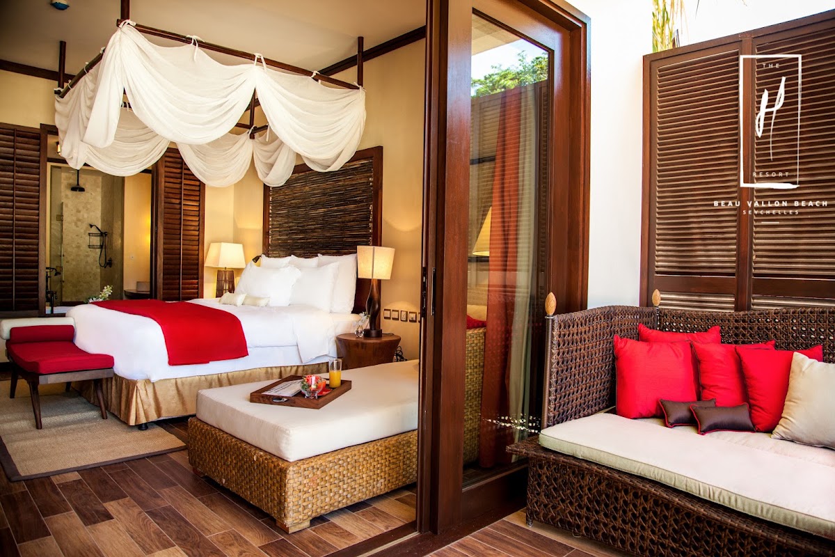 STORY Seychelles guestroom with canopy bed draped in white fabric, red bedding, woven lounge seating with red pillows, and dark wood furnishings.