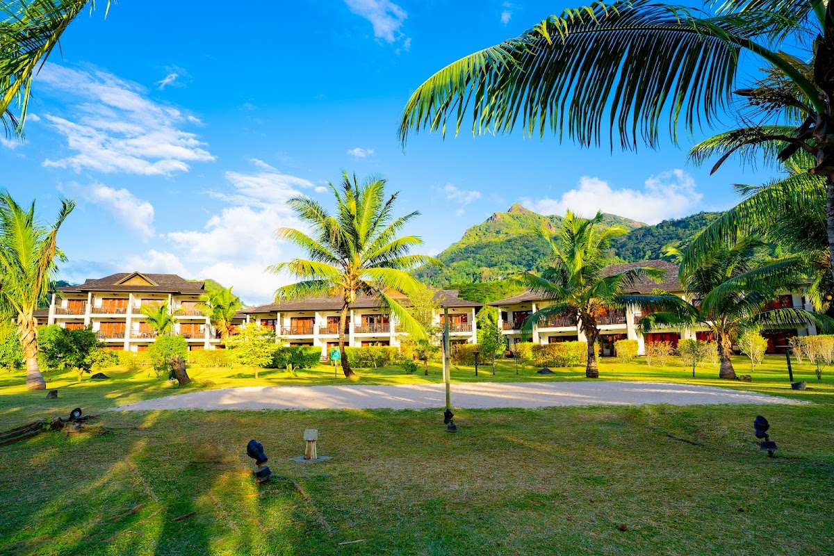 Resort bungalows with white trim and wooden accents line a manicured lawn dotted with palm trees, with forested mountains rising behind under blue sky.