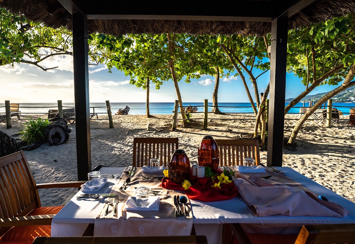 Beachfront dining setup under a thatched-roof pavilion at Story Seychelles, with set table, ocean view, and palm trees framing the sandy shore.