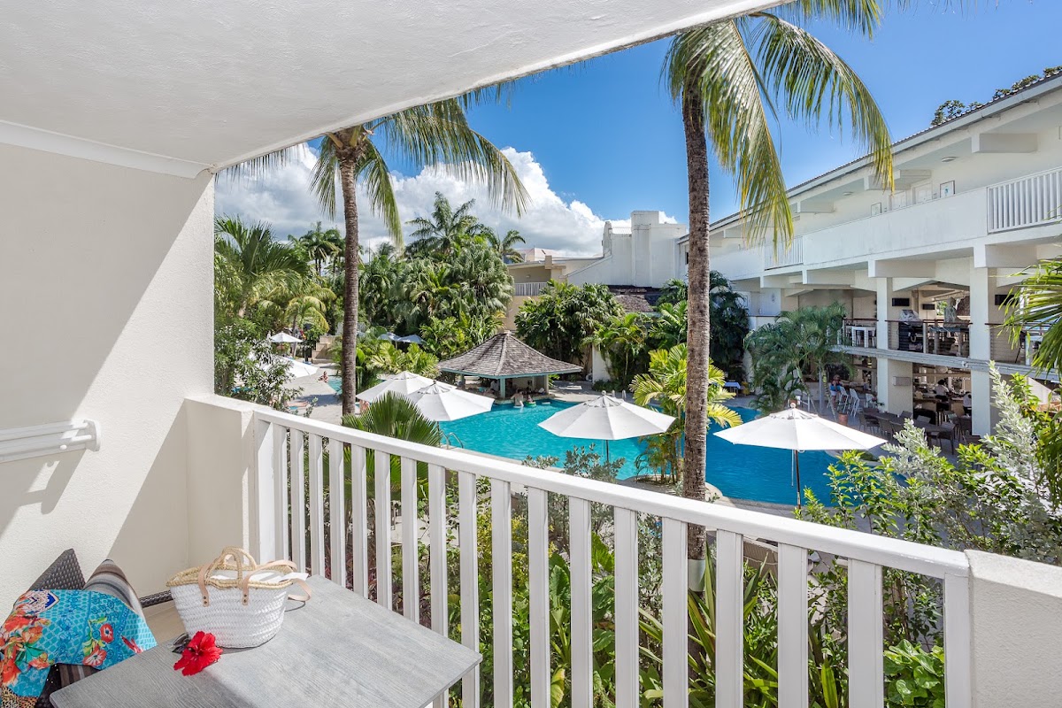 View from a room balcony overlooking the pool area with white umbrellas, palm trees, and multi-story white resort buildings at Sugar Bay Barbados.