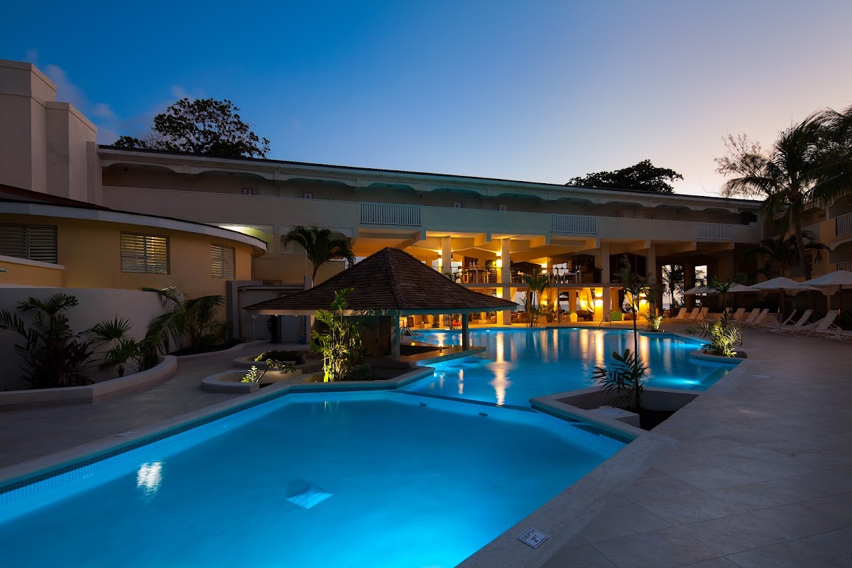 Multiple illuminated pools with a central gazebo at twilight, resort buildings and palm trees visible beyond.